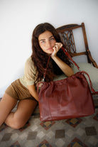 Woman sitting on a chair holding a burgundy leather bag. Elliot bag neuville