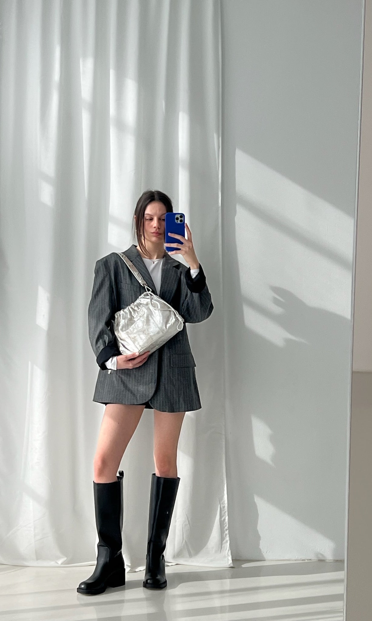 A person holding a small, gold-colored, genuine leather Elliot Mini handbag with a shoulder strap, in front of a mirror.