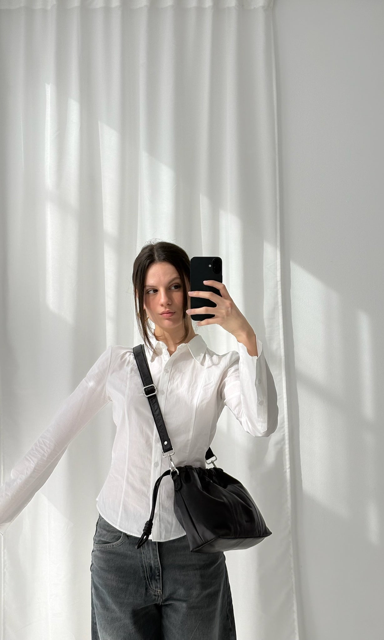 A person wearing a white shirt and gray pants is holding a small black leather bag with a shoulder strap over their shoulder, standing in front of a window with white curtains.
