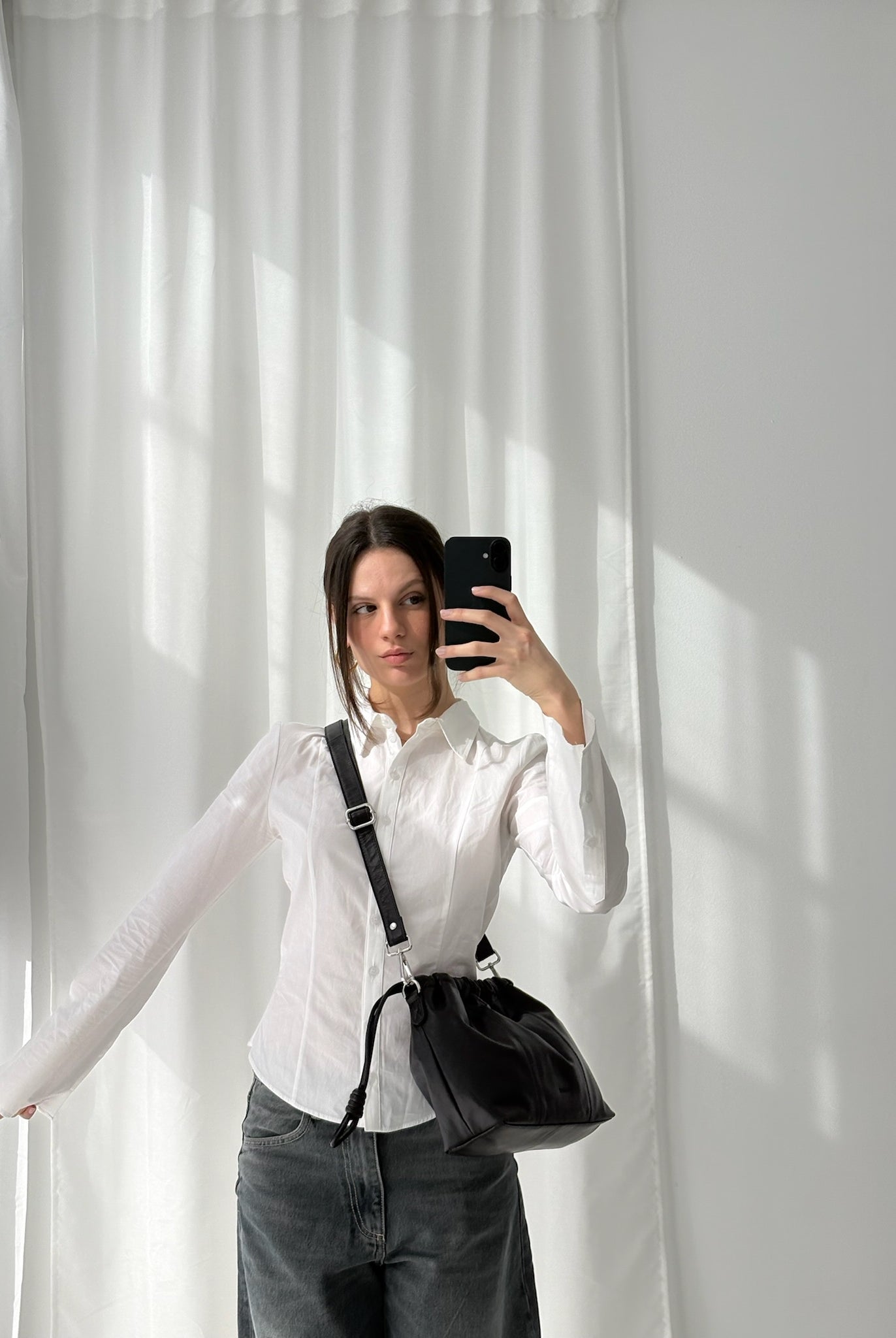 A person wearing a white shirt and gray pants is holding a small black leather bag with a shoulder strap over their shoulder, standing in front of a window with white curtains.