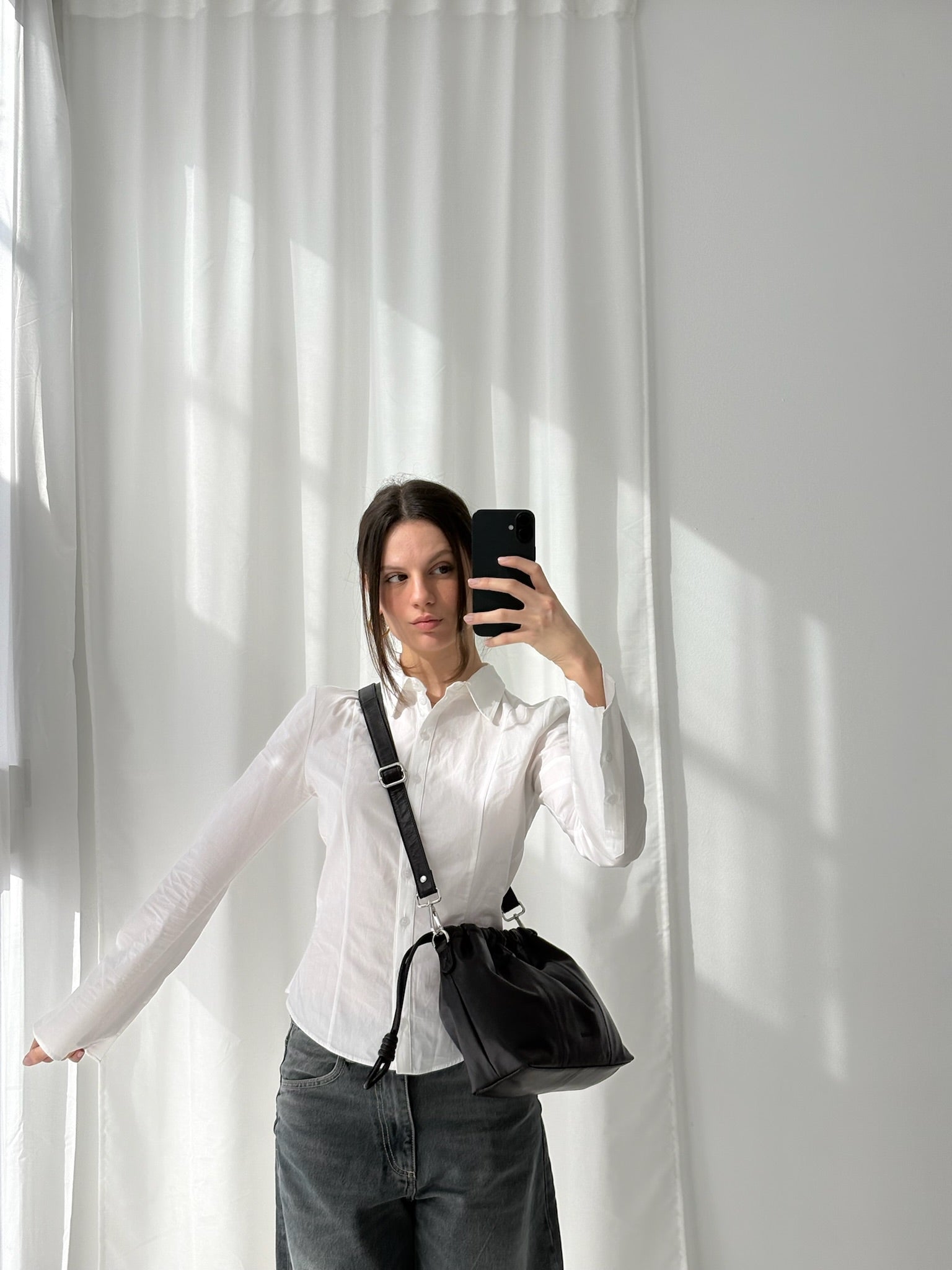 A person wearing a white shirt and gray pants is holding a small black leather bag with a shoulder strap over their shoulder, standing in front of a window with white curtains.