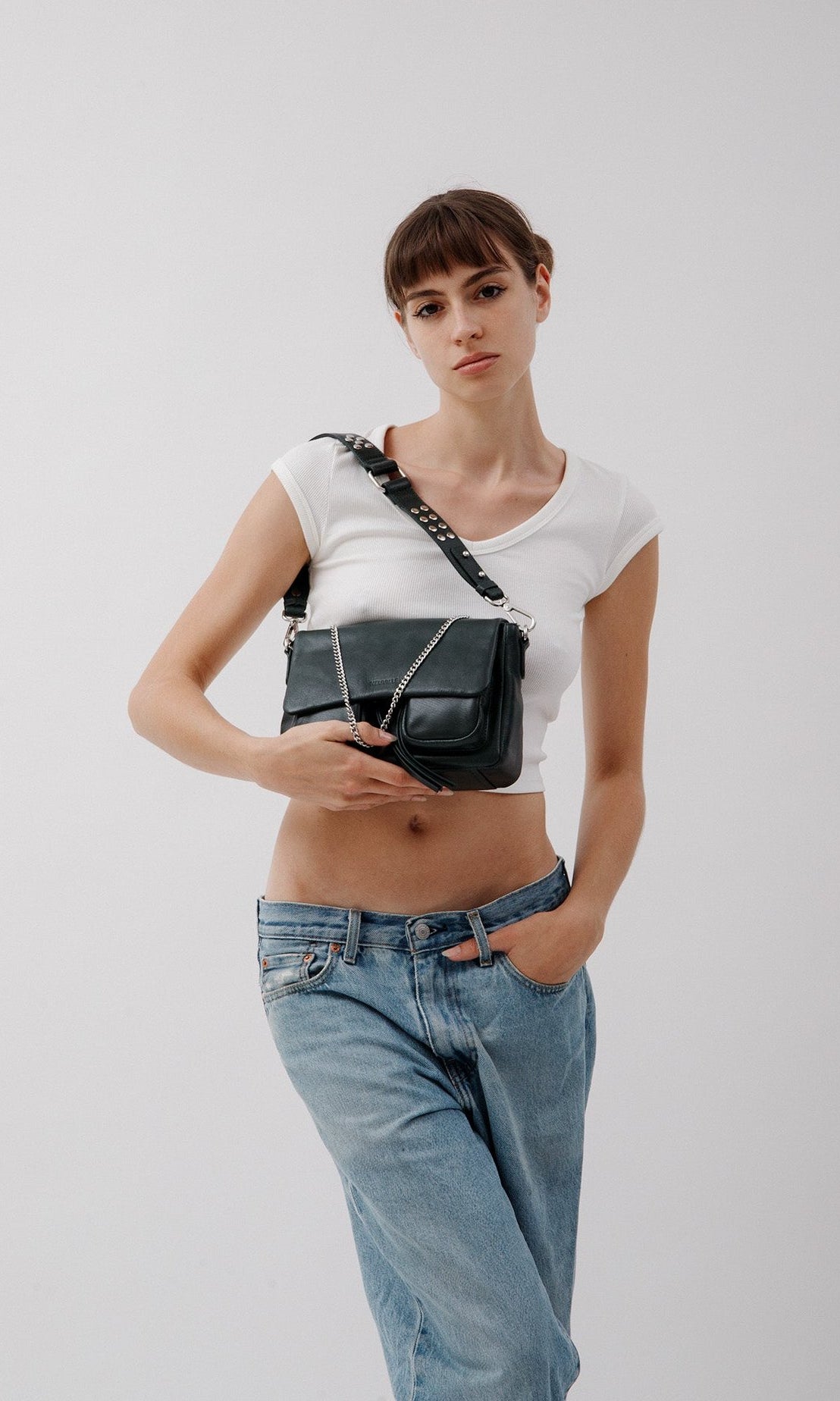 A woman wearing a white tank top and blue jeans, holding a Aqua Maxke leather crossbody bag with a chain strap over her shoulder.