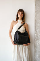 Woman holding a black  leather Neuville bag against a white wall. Elliot bag Neuville