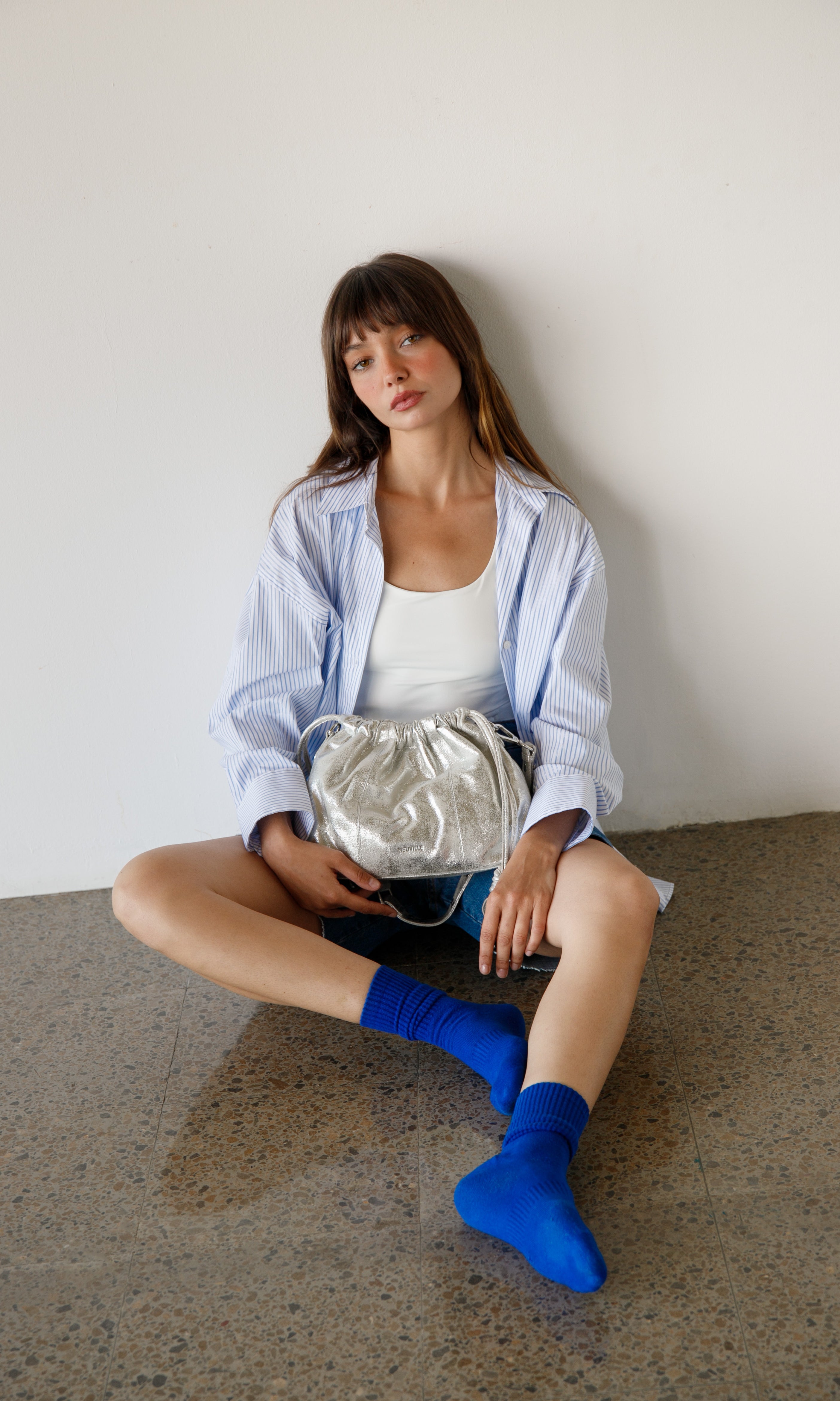 Woman sitting on a concrete floor holding a silver handbag against a white wall.