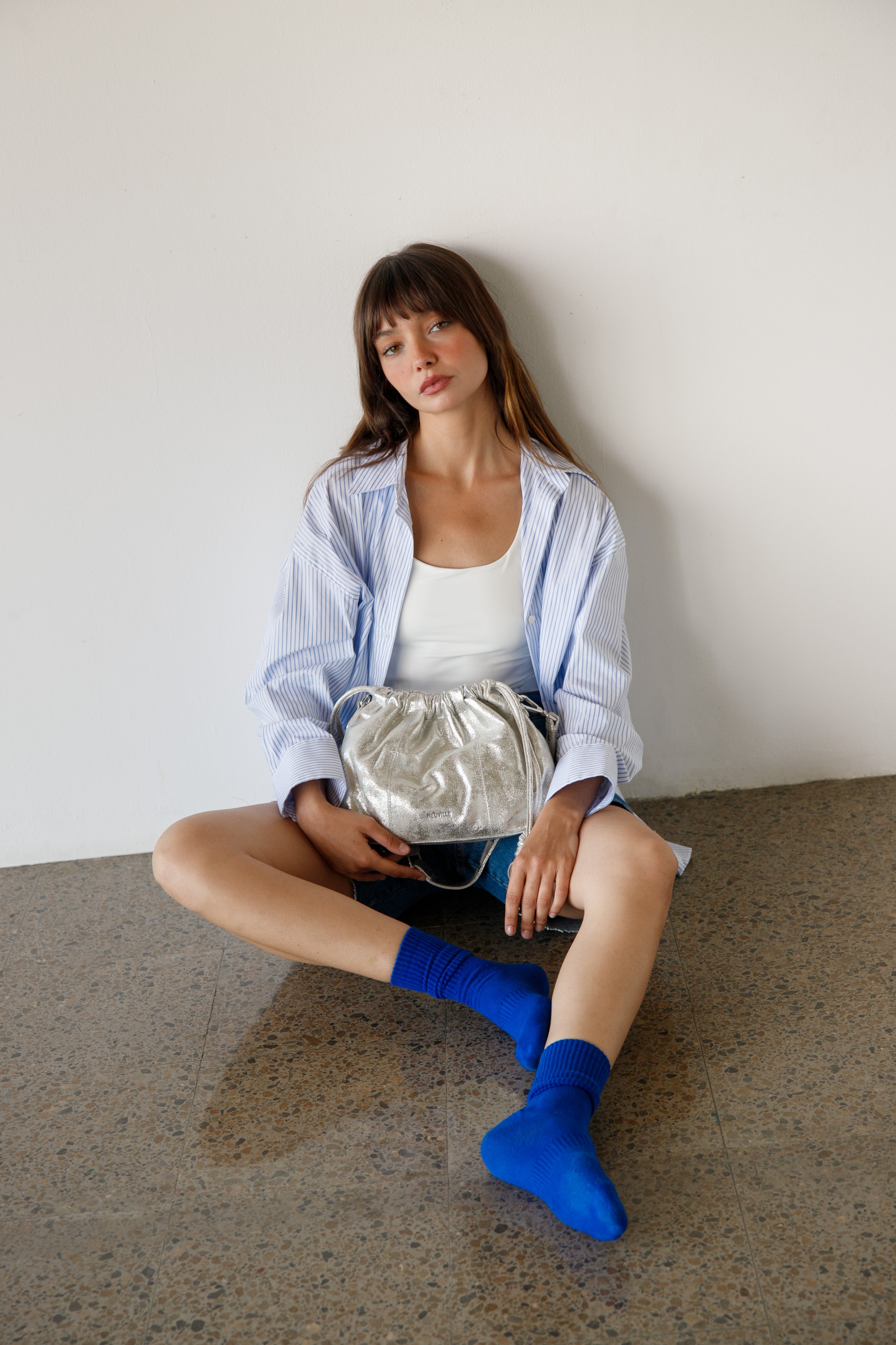 Woman sitting on a concrete floor holding a silver handbag against a white wall.
