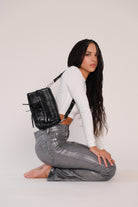 A person wearing a white top and gray trousers, kneeling and posing with a black leather handbag featuring a shoulder strap.