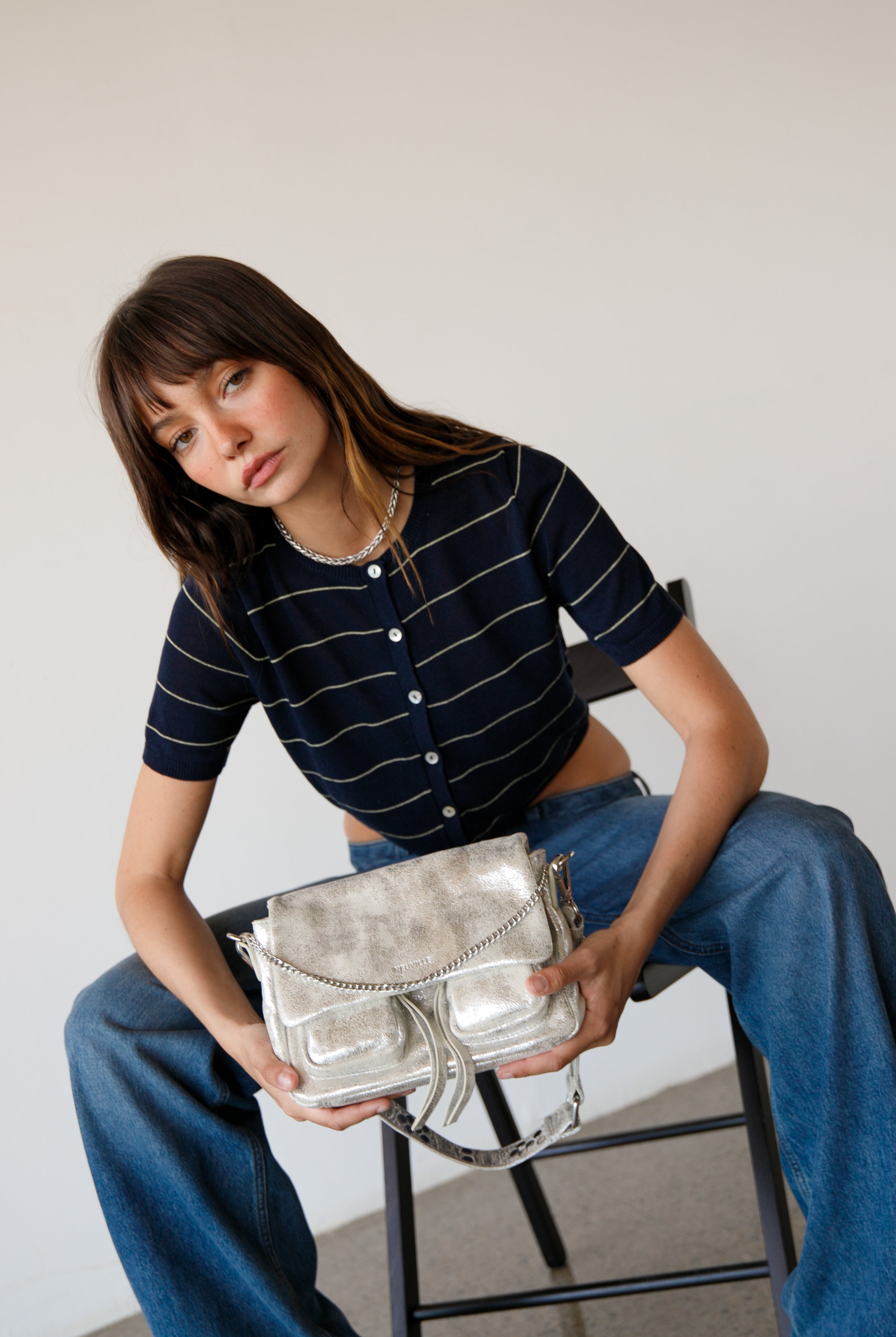 Woman holding a silver handbag while sitting on a chair against a plain background. wearing maxke sand gold leather shoulder bag of the brand neuville
