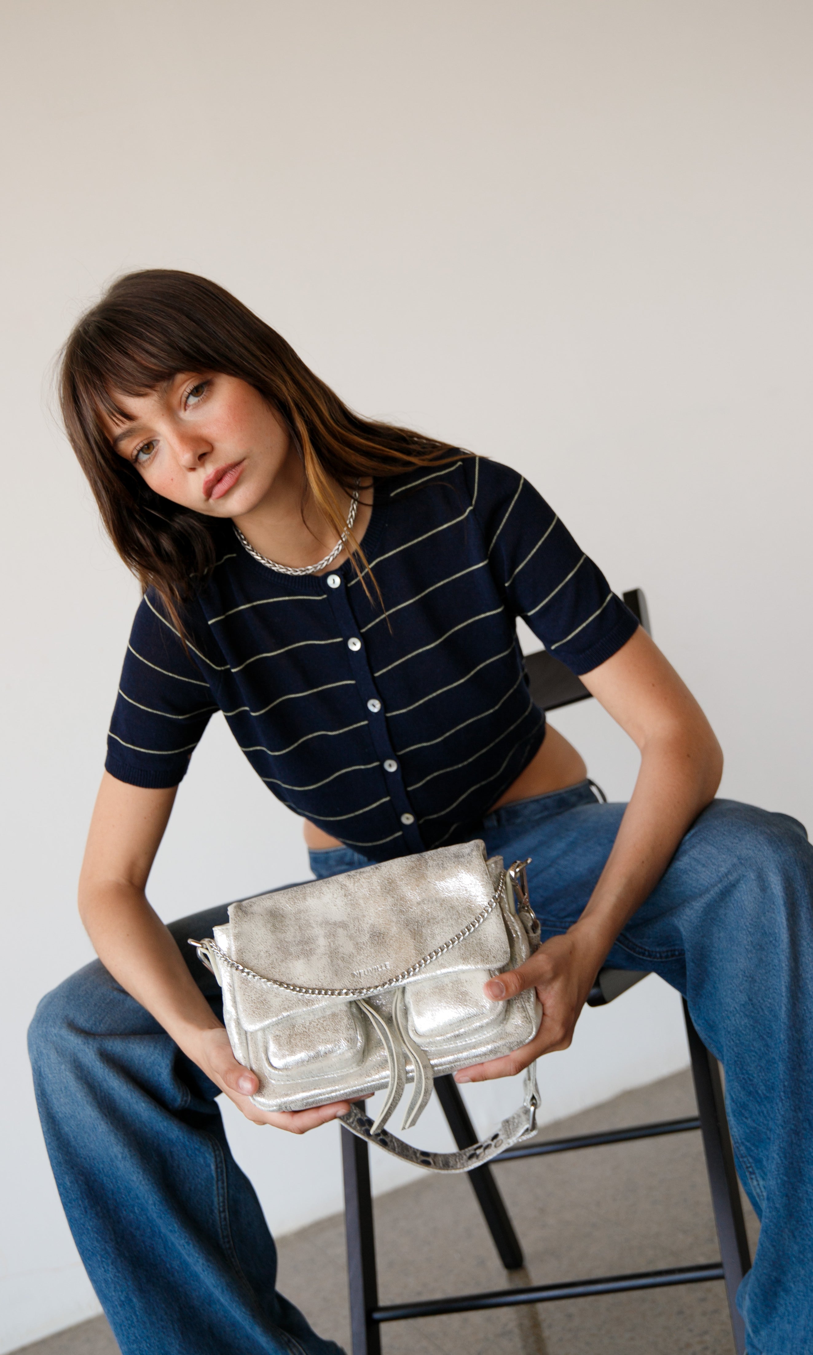 Woman holding a silver handbag while sitting on a chair against a plain background. wearing maxke sand gold leather shoulder bag of the brand neuville