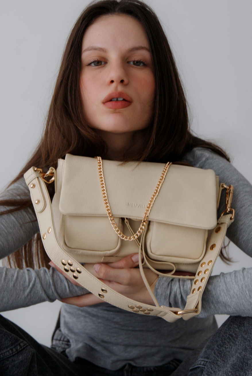A person holding a medium-sized beige Maxke bag with gold hardware. The bag features a leather shoulder strap, a crossbody strap, and a chain shoulder strap, as well as external zippered pockets.