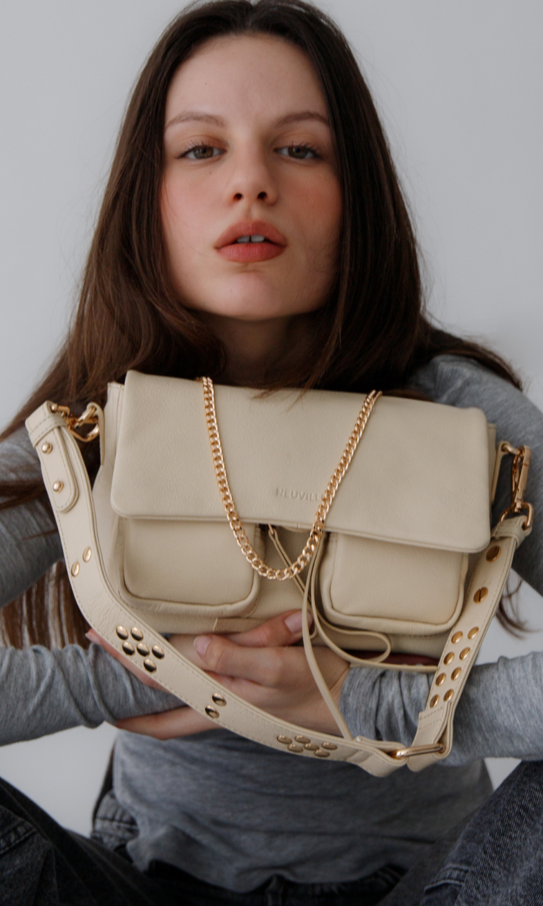 A person holding a medium-sized beige Maxke bag with gold hardware. The bag features a leather shoulder strap, a crossbody strap, and a chain shoulder strap, as well as external zippered pockets.