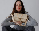 A person holding a medium-sized beige Maxke bag with gold hardware. The bag features a leather shoulder strap, a crossbody strap, and a chain shoulder strap, as well as external zippered pockets.