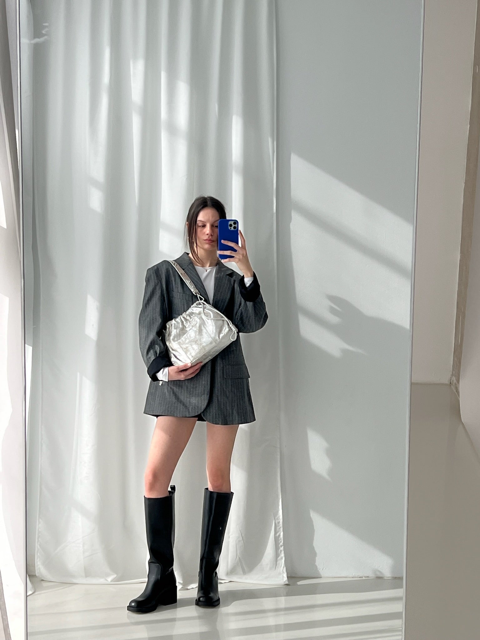 A person holding a small, gold-colored, genuine leather Elliot Mini handbag with a shoulder strap, in front of a mirror.