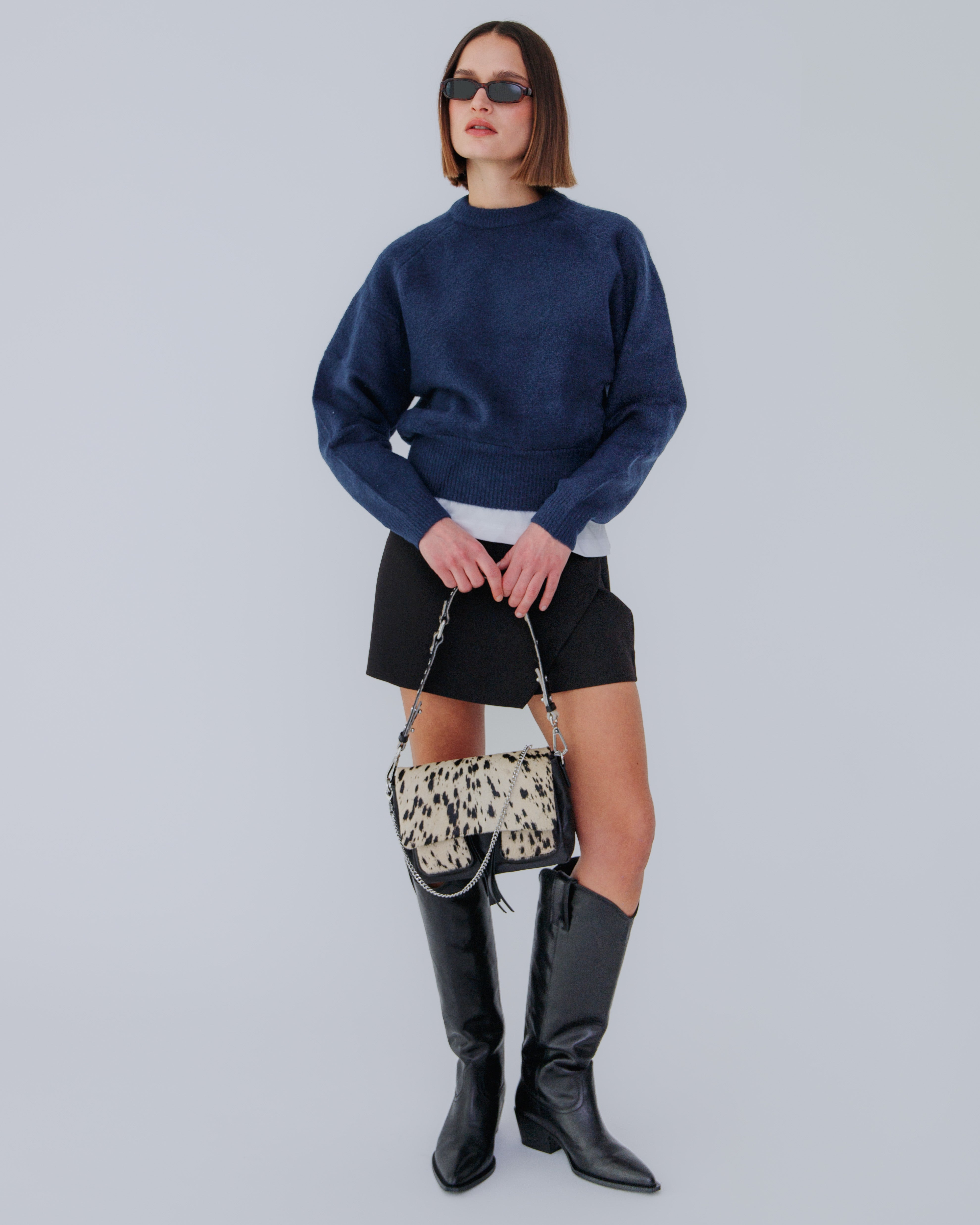 A person wearing a blue sweater and black skirt, holding a black and white patterned handbag with a shoulder strap.