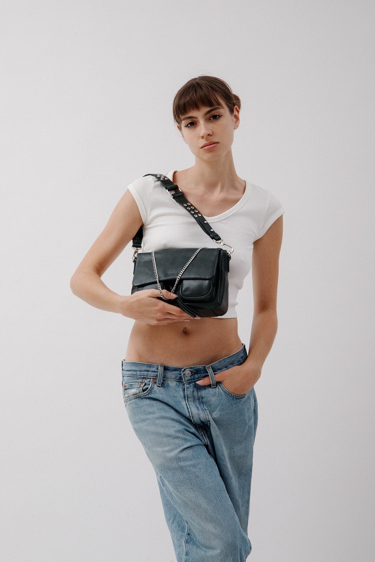 A woman wearing a white tank top and blue jeans, holding a Aqua Maxke leather crossbody bag with a chain strap over her shoulder.