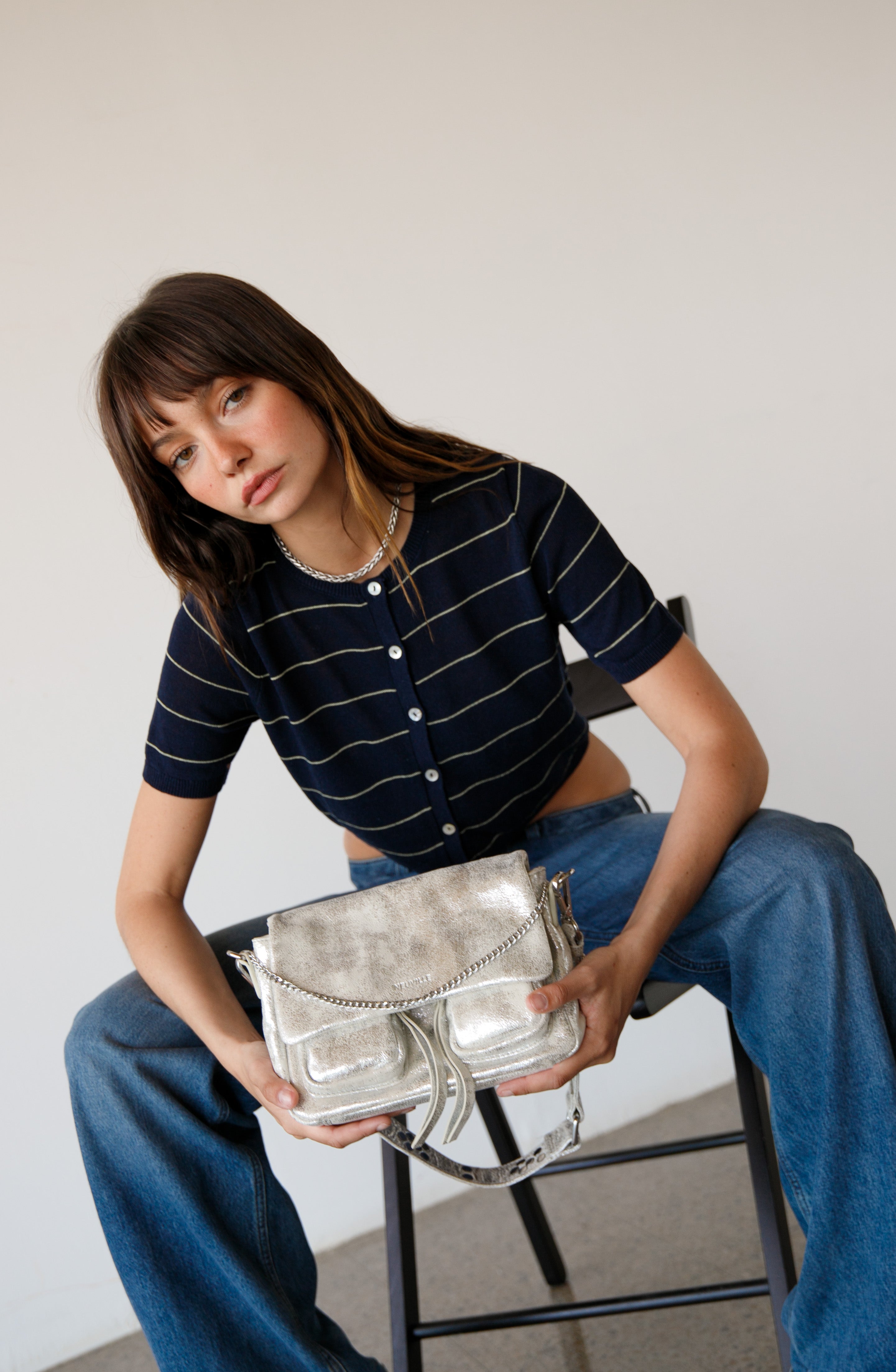 Woman holding a silver handbag while sitting on a chair against a plain background. wearing maxke sand gold leather shoulder bag of the brand neuville